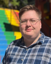 A photo of Morelle Maddox in front of a rainbow painted staircase, wearing a blue flannel shirt and metal framed glasses. 